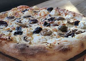 Pizza Bianca mit Oliven und Pilzen Eine ausgebackene Pizza Bianca mit Ricotta, Oliven und Pilzen liegt auf einem Holzbrett.