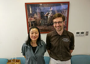 Tomoko, Übersetzerin und Organisatorin der Reise, und ich. Lutz Geißler und Tomoko stehen vor einem gerahmten Bild mit Holzofenmotiv.