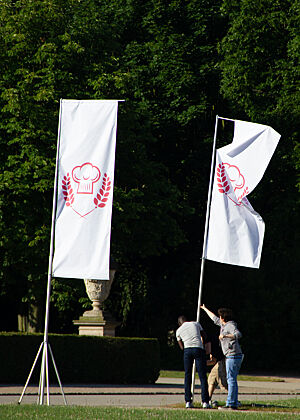 Weiße Fahnen mit dem rotem Emblem der Sendung „Deutschlands bester Bäcker“ werden aufgestellt.