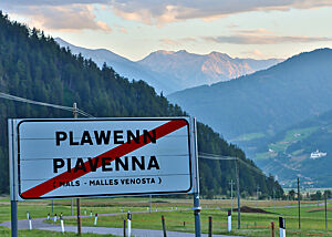 Wir verabschieden uns von Plawenn. Auf dem Ortsschild von Plawenn steht der Ortsname auf Deutsch und Italienisch. Im Hintergrund steigen die bewaldeten Hänge zu grauen Felsen auf.