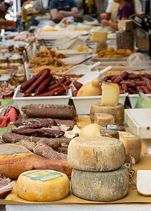 Auf den Marktständen liegt eine reiche Auswahl an runden Käselaiben und roten, getrockneten Würsten.