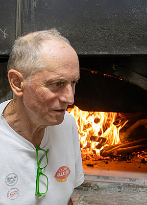 Holzofenbäcker Giovanni Larghetti vor einem seiner Öfen. Giovanni steht vor einem seiner Öfen, in dem durch die geöffnete Klappe der rote Schein des Feuers sichtbar ist.