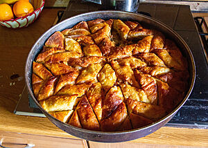 Eine albanische Süßspeise mit Einschnitten in Form einer Schneeflocke liegt goldbraun ausgebacken in einer runden Backform.