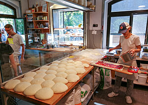 Auf der Arbeitsfläche der Backstube liegen Brot-Teiglinge; ein Pizzateigling wird mit Tomatensoße bestrichen.
