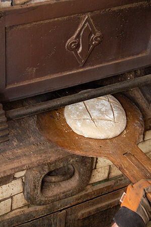 Ein runder Brotteigling wird mit einem Holzschieber in den heißen Ofen geschoben.