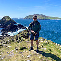 Lutz Geißler steht in Wander-Outfit mit Kamera auf den Sandsteinen des Dunmore Head, umgeben von azurblauem Wasser.