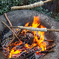 In einer Feuerschale liegen brennende Äste und Glut. Darüber werden zwei Äste gehalten, die an einem Ende mit Stockbrotteig umwickelt wurden.