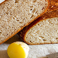 Ein aufgeschnittener Laib Brot mit dunkelbrauner, glänzender Kruste und heller, luftiger Krume liegt zusammen mit einer geschälten, gekochten Kartoffel und einem Brotmesser auf einem Leinentuch.