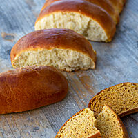 Eine Reihe angeschobener, ovaler Milchbrötchen mit dunkelbraun glänzender, weicher Kruste liegt neben einem Zwieback mit brauner, getrockneter Krume.