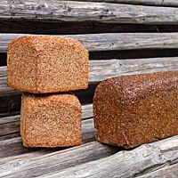 Auf einer rustikalen Holzbank liegen ein echter Pumpernickel, dunkler im Aussehen, nach überlieferter Tradition mit Brandsauerteig hergestellt (rechts) und das gleiche Brot, deutlich heller, mit deutlich kürzerer Backzeit als Roggenschrotbrot/Schwarzbrot (links).