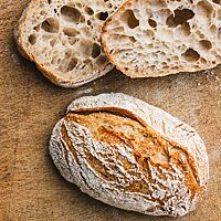 Auf einem Holzuntergrund liegen zwei Weizensauerteigbrötchen. Ein ganzes Brötchen zeigt die goldbraun ausgebackene, bemehlte, krosse Kruste; ein halbiertes Brötchen zeigt die grobe, unregelmäßige Porung der saftigen Krume.