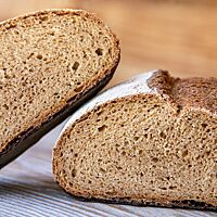 Auf einem gräulichen Holzuntergrund liegt ein halbiertes Dinkelvollkornsauerteigbrot mit einem Altbrot-Brühstück. Die eine Brothälfte liegt seitlich auf die untere Hälfte gelehnt. Der Anschnitt zeigt die lockere, elastische, kleinporige Krume.