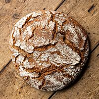 Ein rundes, rustikal aufgerissenes Dinkelbrot mit bemehlter Kruste liegt auf einem rustikalen Holzuntergrund.