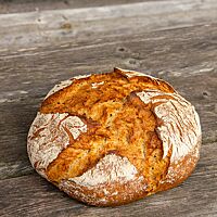 Ein rustikal aufgerissenes, goldbraun ausgebackenes, rundes Brot mit bemehlter Kruste liegt auf einem Holztisch. 