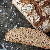 Auf einem dunklen Steinuntergrund liegt ein angeschnittenes Schwarzwälder Brot mit bemehlter, rustikal aufgerissener Kruste. Im Vordergrund liegt eine Scheibe des saftigen, lockeren Brotes.