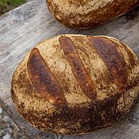 Ein dunkel ausgebackenes, dreimal quer eingeschnittenes, ovales Brot mit bemehlter Kruste liegt auf einem verwitterten Holztisch.