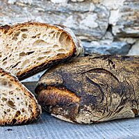 Ein kräftig ausgebackenes Weißbrot mit dunkler, bemehlter Kruste liegt auf einem Holzuntergrund vor einer Natursteinmauer.