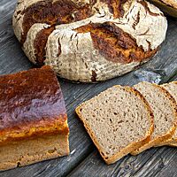 Auf einem gräulichen Holzuntergrund liegen ein doppeltgebackenes Bauernbrot und ein Kastenlandbrot vor dem drei Scheiben des Brotes liegen. Das Kastenbrot hat eine glänzende, glatte Kruste, das Bauernbrot hat eine dunkel ausgebackene, rustikal aufgerissene, bemehlte Kruste.