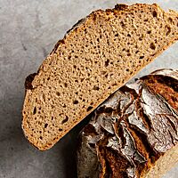 Ein halbiertes, rustikal aufgerissenes Roggenbrot mit bemehlter Kruste liegt auf einem grauen Untergrund. 