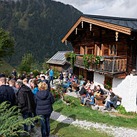 Menschen stehen oder sitzen vor der Almhütte in der Sonne.