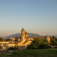 Die Stadt Urbino strahlt im letzten Abendlicht. In der Mitte erhebt sich ein Kirchturm und im Hintergrund sieht man grüne Hügel.