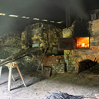 Die zwei baugleichen Holzöfen stehen nebeneinander in einer dunklen Scheune. Im linken Ofen ist dank der offenen Ofentüre das Leuchten des Feuers sichtbar.