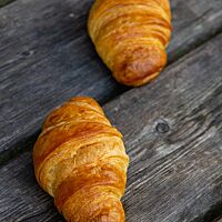 Zwei goldbraun ausgebackene Croissants aus hellem Mehl liegen auf einem rustikalen Holztisch.