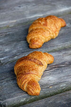 Croissants (Vollkorn und hell). Zwei goldbraun ausgebackene Vollkorncroissants liegen auf einem rustikalen Holztisch.