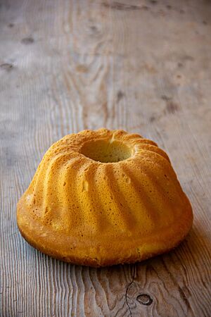 Almhupf. Der goldbraun ausgebackene, aus der Form gestürzte Guglhupf liegt auf einem rustikalen Holztisch.