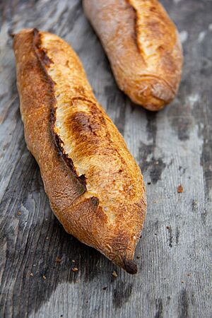Baguette au Levain (2020). Zwei kräftig ausgebackene Baguettes, rustikal aufgebrochen auf einer rustikalen Holzunterlage.