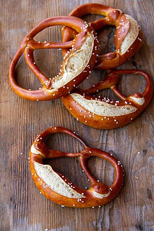 Belaugte Schnee-Brezn. Vier an den Brezelbäuchen eingeschnittene Laugenbrezeln mit Salz auf der Kruste liegen auf einem rustikalen Holztisch.