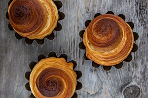 Drei in Brioche-Formen gebackene, aufgerollte Brioche liegen auf einem rustikalen Holztisch.