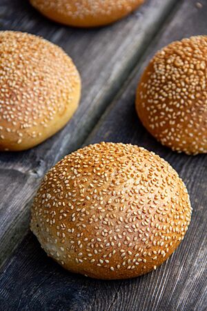 Vier goldbraun ausgebackene, in Sesam gewälzte Briochebrötchen liegen auf einem rustikalen Holztisch.