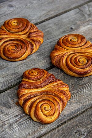 Drei flache Franzbrötchen mit schöner Schneckenform und glänzender Zimt-Zucker-Füllung liegen auf einem grauen Holztisch.