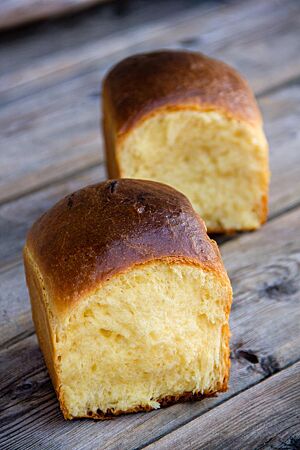 Zwei würfelförmige Brioche liegen auf einem Holztisch und zeigen die glänzende, glatte Kruste und die wattige hellgelbe Krume.