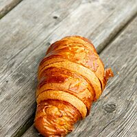 Ein goldbraun ausgebackenes Croissant mit glänzender, blättriger Kruste liegt auf einem grauen Holztisch.