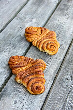 Zwei goldbraun ausgebackene, flache Franzbrötchen mit einer sichtbaren Füllung aus Zimt und Zucker liegen auf einem Holztisch.