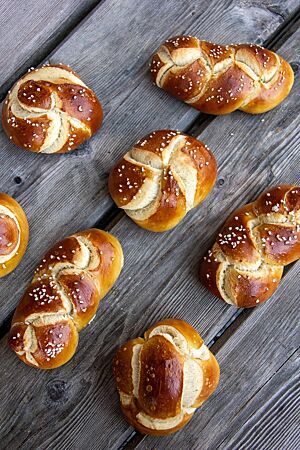 Mehrere, braun glänzende Laugenbrötchen, als Zopf oder Semmel geformt, liegen auf einem grauen Holztisch.