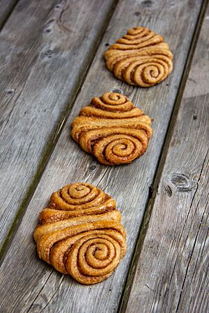 Drei flache Franzbrötchen aus Vollkornteig mit einer Zimtzucker-Füllung liegen auf einem grauen Holztisch.