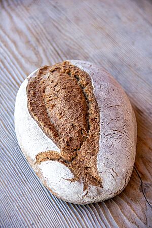 Das Dinkelvollkorn-Sauerteigbrot mit mittig aufgerissener, bemehlter Kruste liegt auf einem hellen Holztisch.