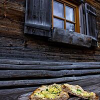 Eine Dinnete liegt auf einer hölzernen Brotschaufel mit langem Griff vor einer Alpenhütte aus dunkel verwitterten Holzbalken.