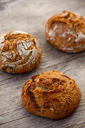 Dreikornbrötchen mit Saaten. Zwei Brötchen mit bemehlter Kruste und rustikalem Ausbund und ein unbemehltes Brötchen liegen auf einem Holztisch.