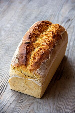 Ein goldbraun ausgebackenes Kastenbrot mit hellen Seiten und mittig aufgerissener Kruste auf der Oberseite liegt auf einem grauen Holztisch.