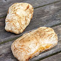 Zwei kleine, goldgelb ausgebackene Ciabattas mit bemehlter Kruste liegen auf einem rustikalen Holztisch.