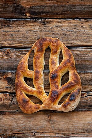 Rauriser Fougasse. Die kräftig ausgebackene, flache Rauriser Fougasse mit dunkelbraunen Stellen auf der Kruste liegt auf einem rustikalen Holztisch. Sie ist vor dem Backen mittig und mehrmals seitlich durchstochen worden, sodass sich mehrere „Zweige“ bildeten.
