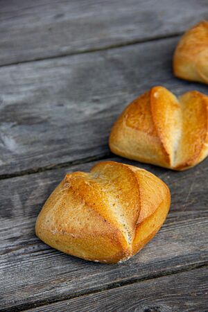 Frühstückssemmeln. Quadratische, goldgelb ausgebacke Brötchen mit tiefem diagonalem Einschnitt liegen auf einem grauen Holztisch.