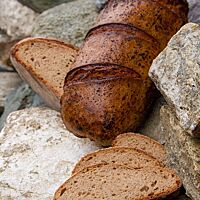 Ein länglicher Laib liegt zwischen grauen Steinen, die einen schönen Kontrast zur kräftig ausgebackenen, leicht glänzenden Kruste des Brotes bieten.