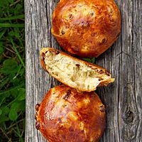 Runde, goldbraune Rosinenbrötchen mit glänzender, dünner Kruste liegen auf einer Holzbank über einem Stück Wiese.