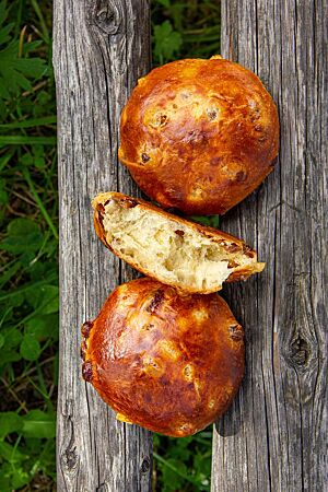 Runde, goldbraune Rosinenbrötchen mit glänzender, dünner Kruste liegen auf einer Holzbank über einem Stück Wiese.