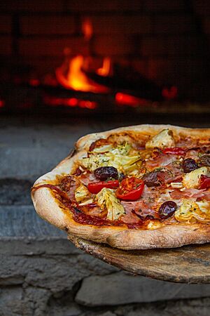 Die dünn belegte Hüttenpizza liegt auf einem Einschießer vor dem Holzbackofen.